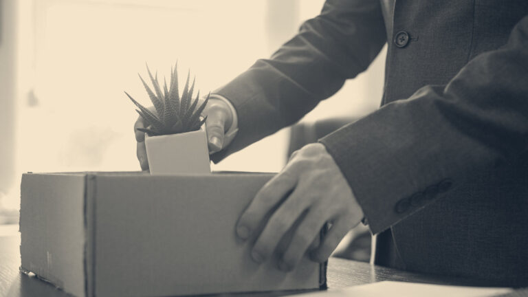A person in a dark suit packs a small potted succulent into a cardboard box on an office desk