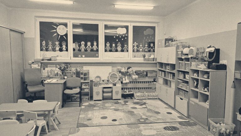 An empty nursery classroom: small yellow chairs at green tables, toy shelves, rugs and children's paper figures taped to the windows