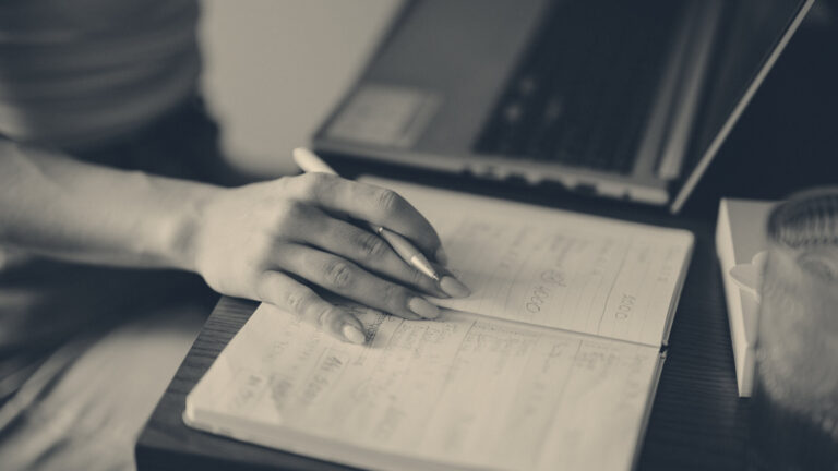 A hand writes notes in a paper diary beside a laptop on a dark wooden desk in soft window light.