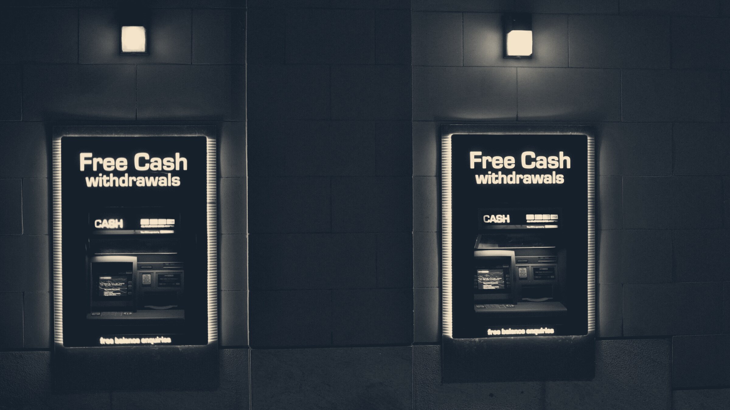 Two blue-lit cash machines recessed in a stone wall at night, sign reading free cash withdrawals