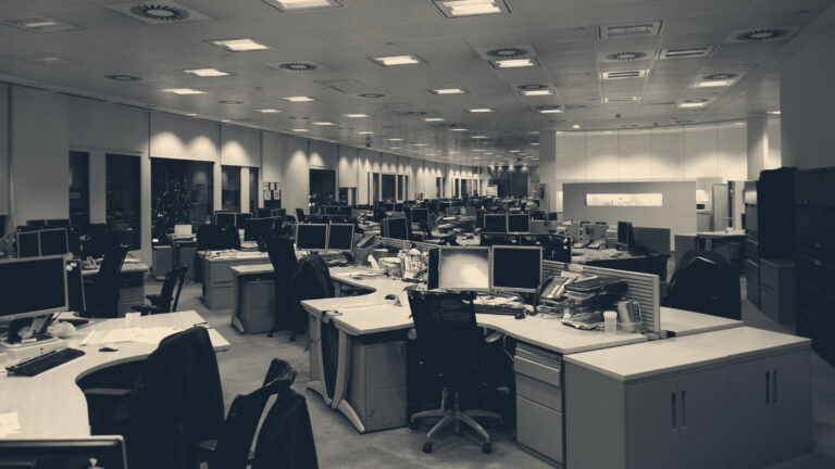 An empty Canary Wharf office floor after dark, lit by ceiling panels and a single glowing monitor on a cluttered desk.