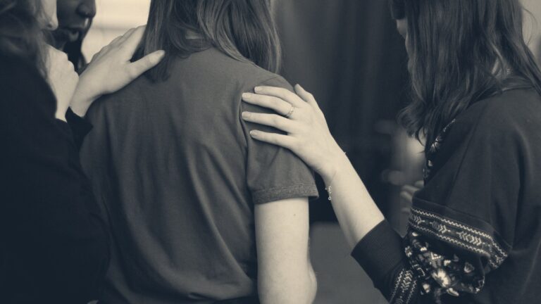 Women stand close in support, hands resting on a colleague's shoulders, in a softly lit room.
