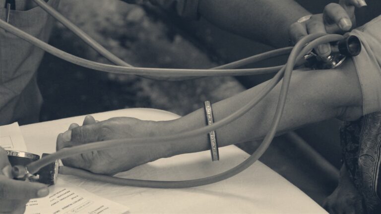 Close-up of a health check: a clinician's hands fit a blood-pressure cuff to a patient's arm over a form