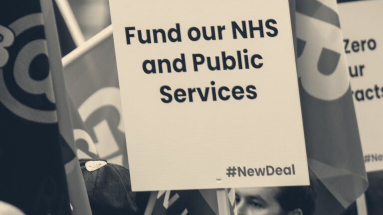 Union placards and flags held aloft at a UK street demonstration; lead placard reads 'Fund our NHS and Public Services'.