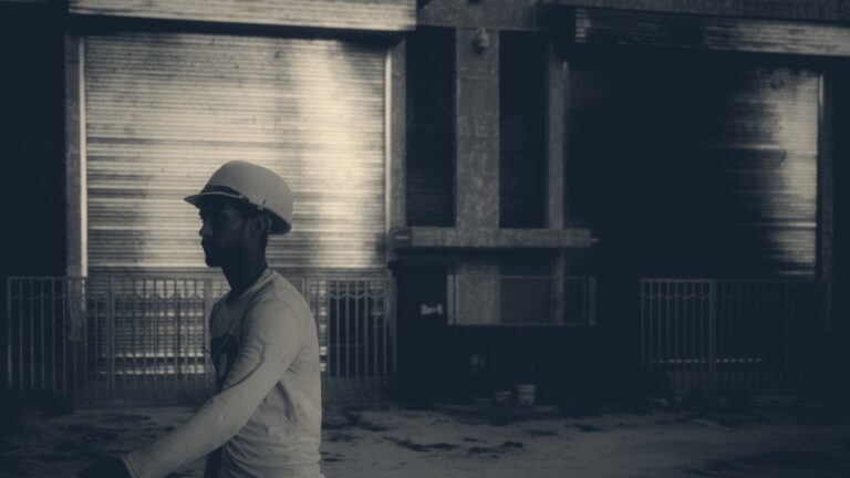 A worker in an orange hard hat walks past shuttered industrial units lit by warm evening light.