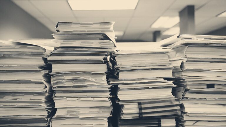 Tall stacks of paper files and folders in a records office under fluorescent ceiling lights.