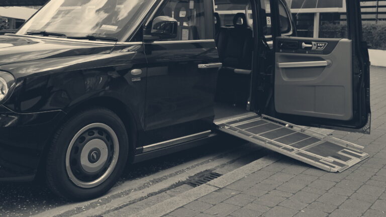 A black London taxi with its side door open and a wheelchair access ramp deployed onto a paved pavement.