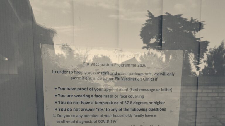 A vaccination rules notice taped to a GP surgery window listing entry conditions, trees reflected in the glass
