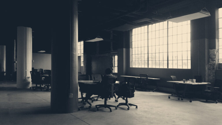 Lone worker at a laptop in a vast dark office, seen from behind, daylight through tall grid windows