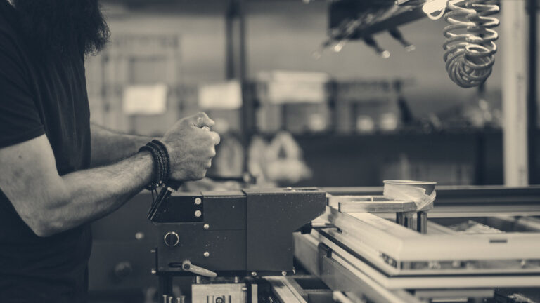 A factory worker operates a conveyor machine, hand reaching toward controls beside a red emergency stop.