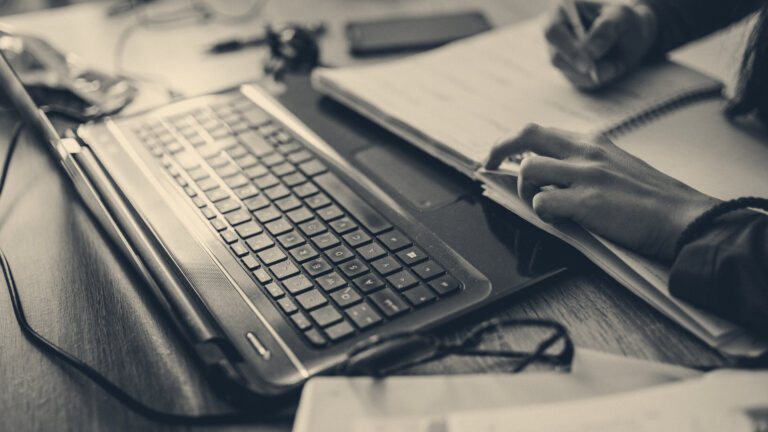 A freelancer writes in a notebook at a desk next to an open laptop, spectacles, and work papers.