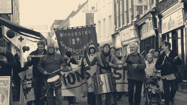 Marchers with drums and a red Worcester Trades Council banner on a UK high street during a pensions demonstration.