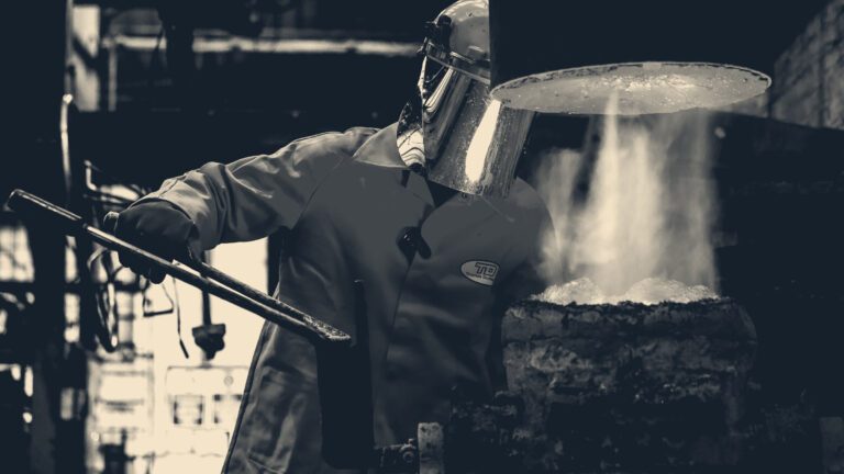 A foundry worker in orange PPE and a raised visor tends molten metal glowing beneath a furnace