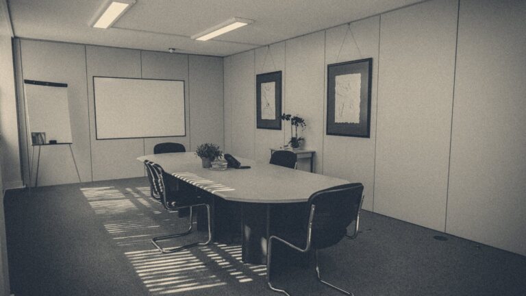 Sunlight through blinds casting stripes across an oval boardroom table, chairs, and whiteboard in an empty UK office.