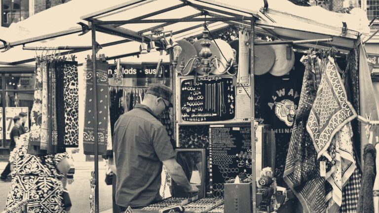 Grey-haired man in a checked cap arranging jewellery on his stall at Cambridge market