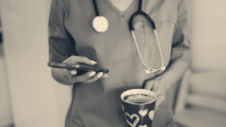 Healthcare worker in teal scrubs with a stethoscope checking a smartphone while holding a red mug in a bright break room