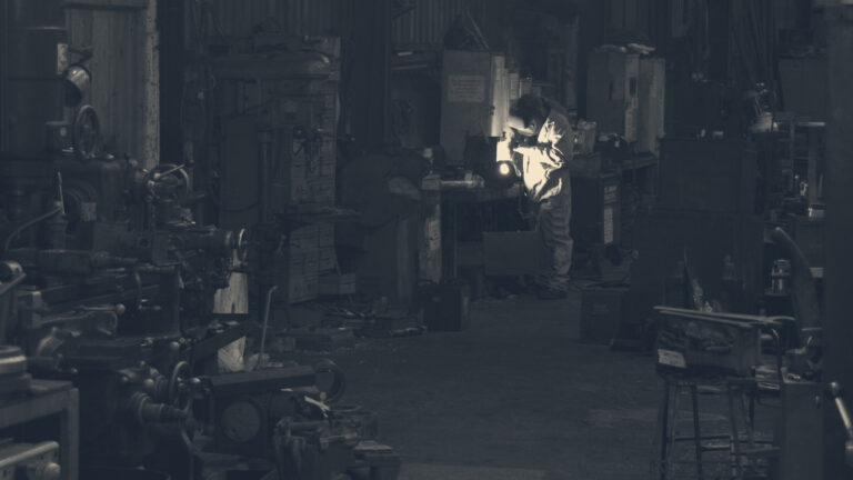 A worker in an orange boiler suit and welding visor welds metal inside a dark Loughborough locomotive workshop.