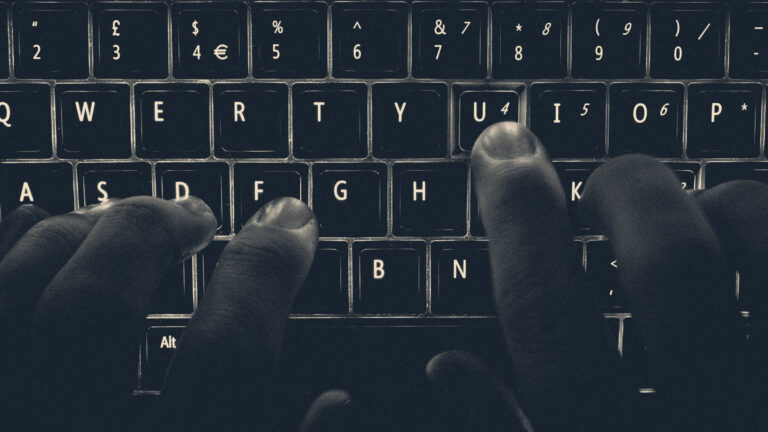 Hands typing on an illuminated UK backlit keyboard in the dark, showing the pound symbol and glowing keys.