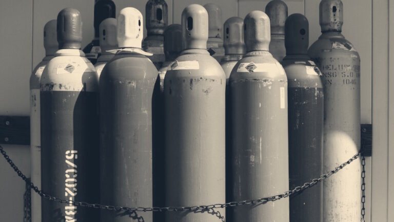 Colour-coded industrial gas cylinders standing upright and chained to a corrugated workshop wall.