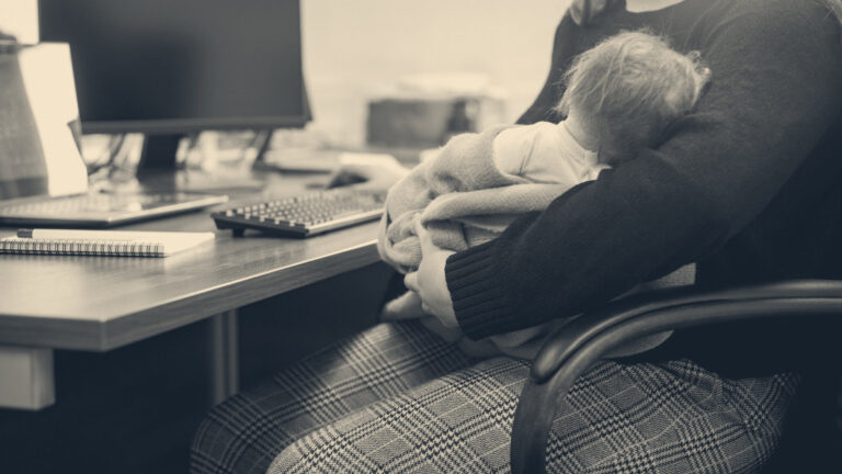 A mother cradles her sleeping newborn at an office desk beside a computer keyboard and monitor during maternity leave