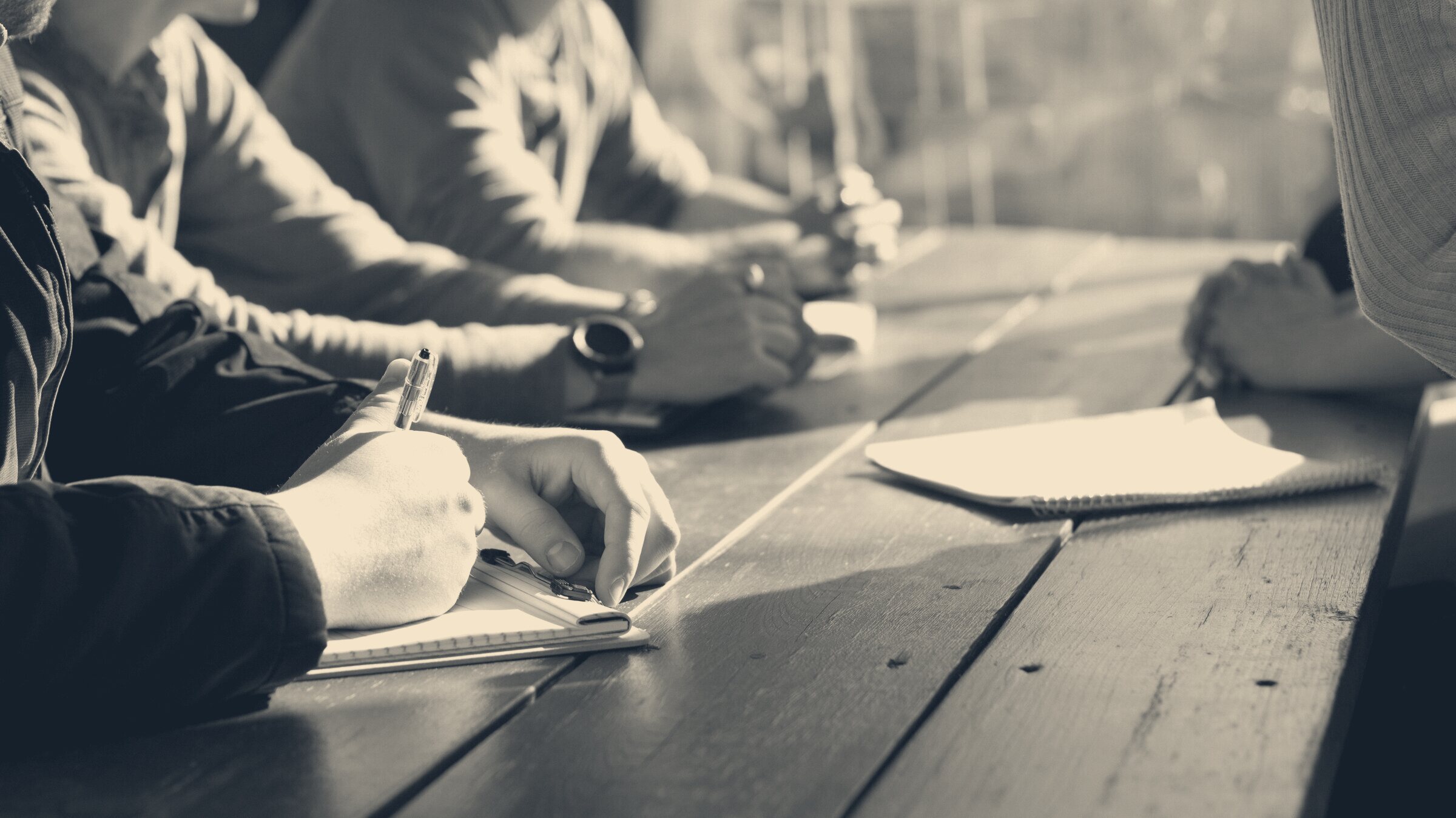 Colleagues sit across a wooden table taking notes on paper pads with pens during a negotiation meeting.