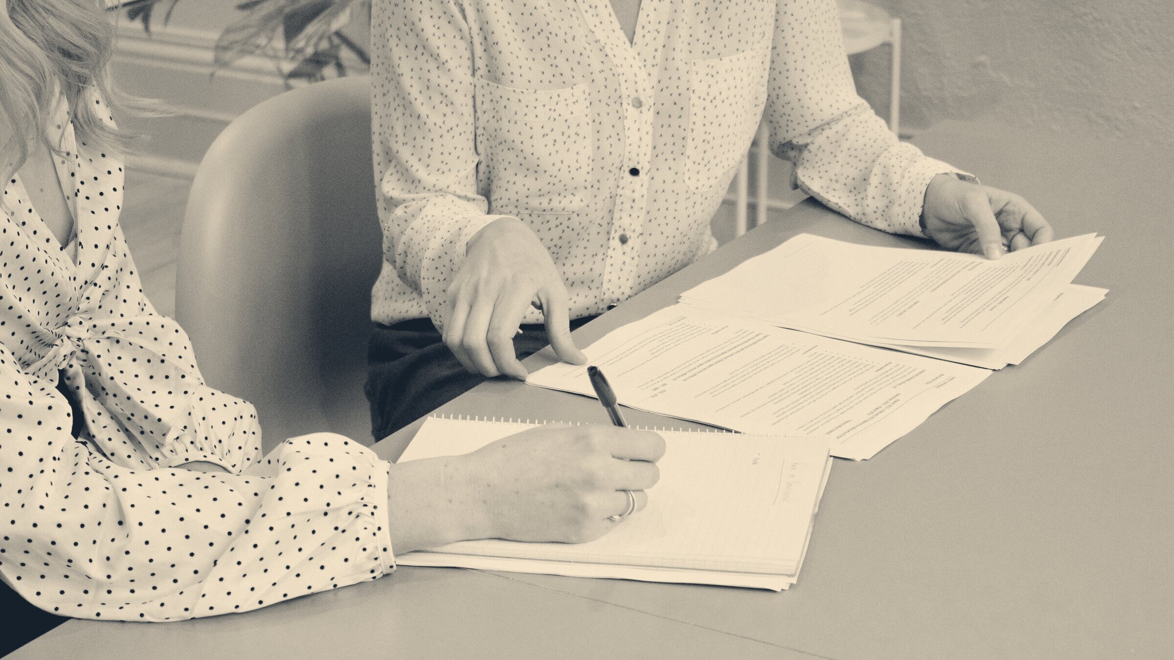 Two employees review printed share scheme documents across an office table, one taking notes with a pen in a spiral pad.