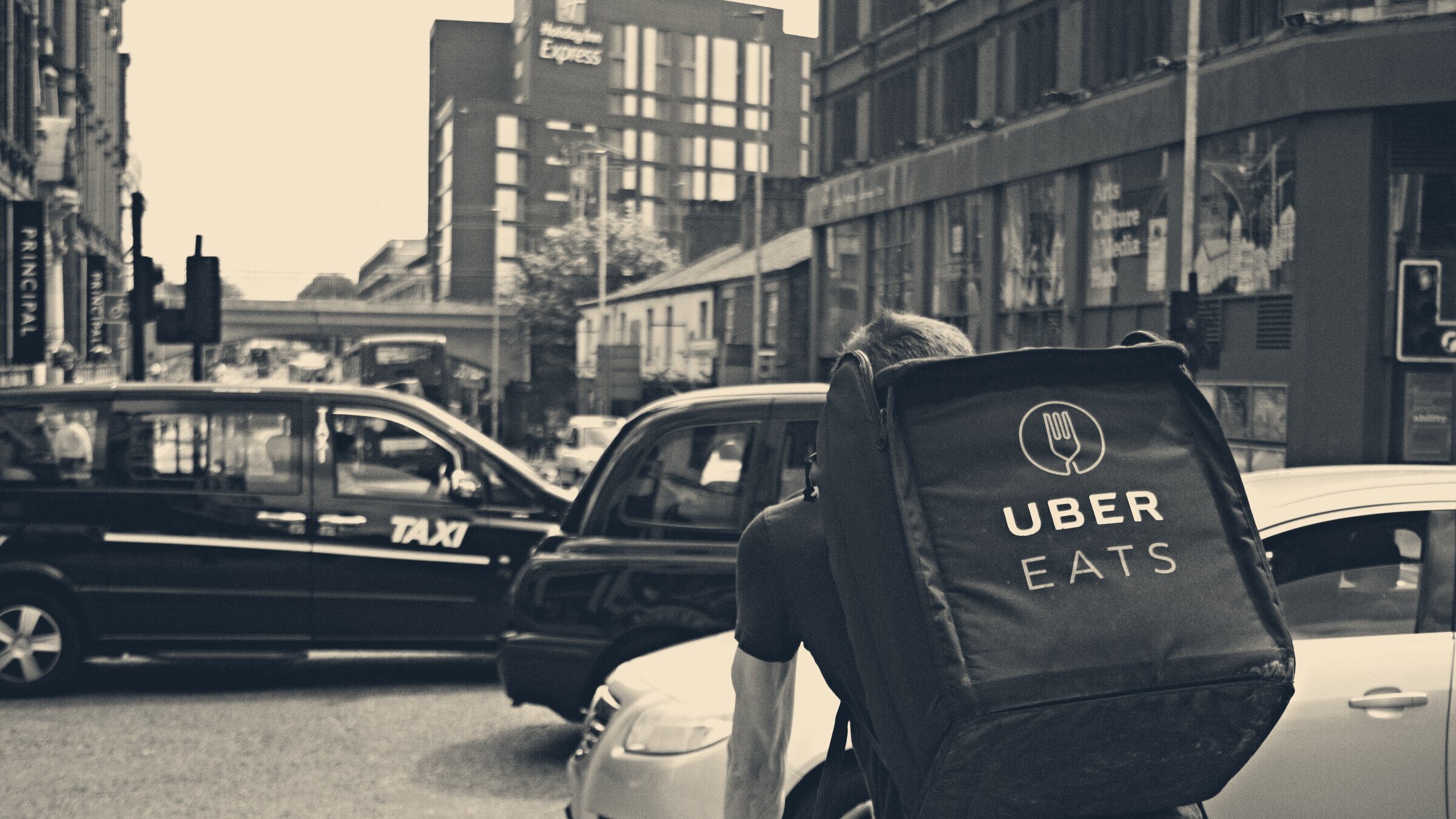 An Uber Eats rider seen from behind, waiting on a bike in Manchester traffic beside a black taxi