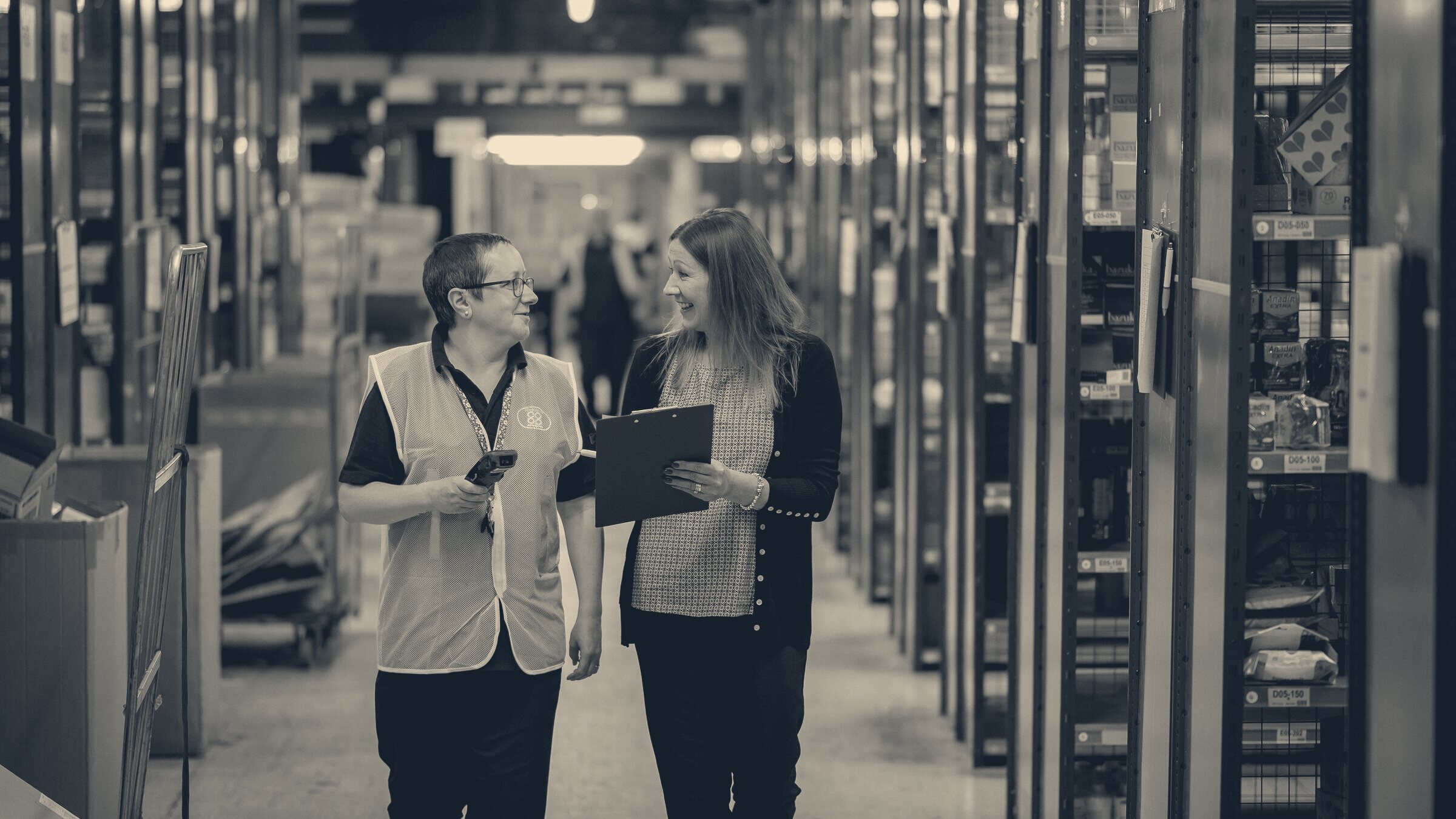 A warehouse worker in a hi-vis vest talks with a colleague holding a clipboard in an aisle of blue racking