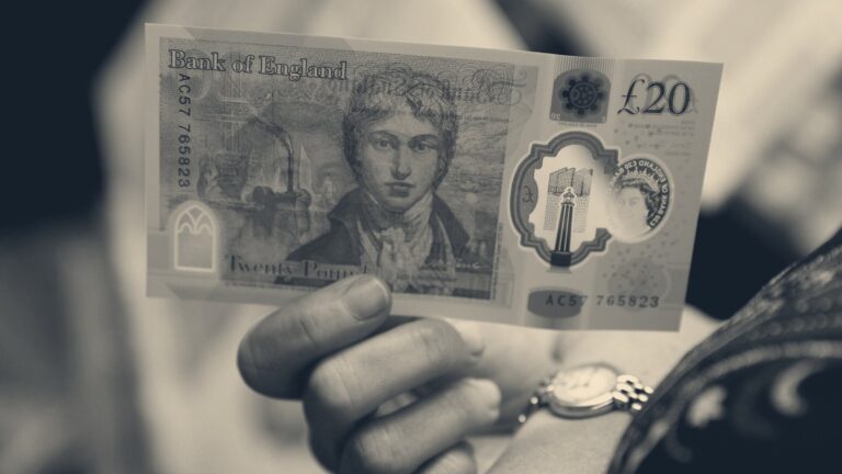 A hand holding a £20 Bank of England note, wristwatch visible, blurred daylight background