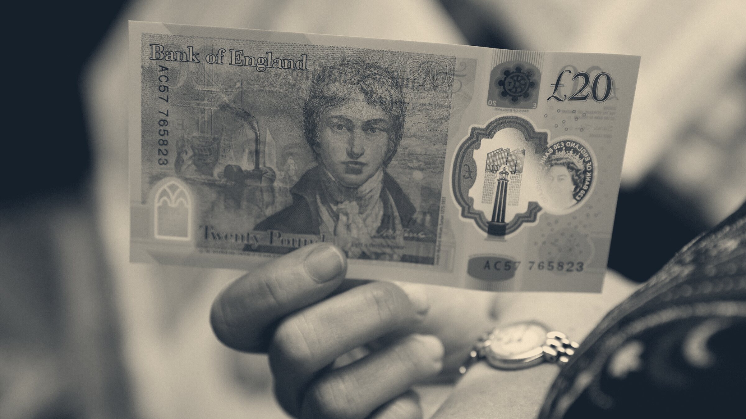 A hand holding a £20 Bank of England note, wristwatch visible, blurred daylight background