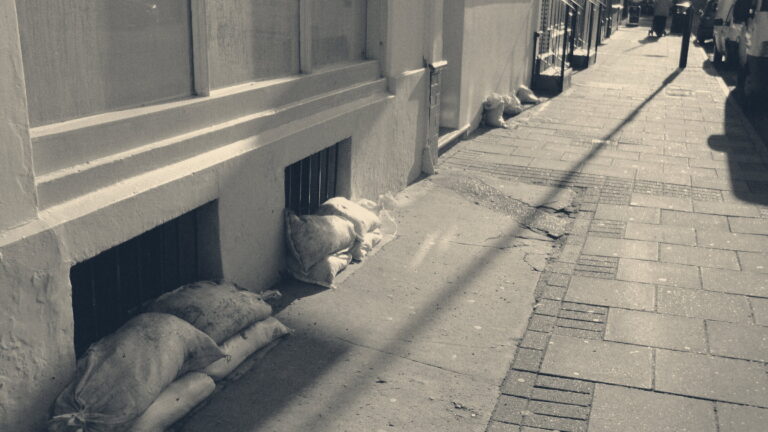 Sandbags piled outside a yellow-painted shopfront and cellar grilles on a sunlit pavement in Ryde, Isle of Wight.
