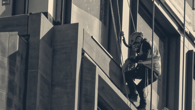 A rope-access worker in an orange helmet and hi-vis vest descends a stone-clad City of London tower on rigged lines.