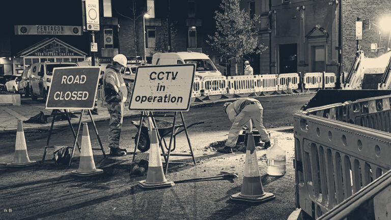 Road workers in orange hi-vis dig and supervise on a cordoned-off Chatham street at night behind cones and barriers.