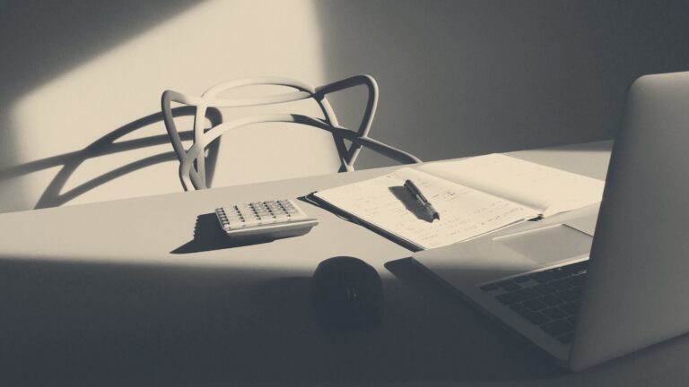 Desk with laptop, desktop calculator, open notebook with notes, and an empty chair casting shadows under sunlight.