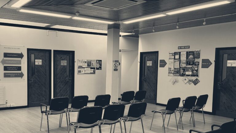 Empty chairs and numbered consulting room doors in a British NHS clinic waiting room