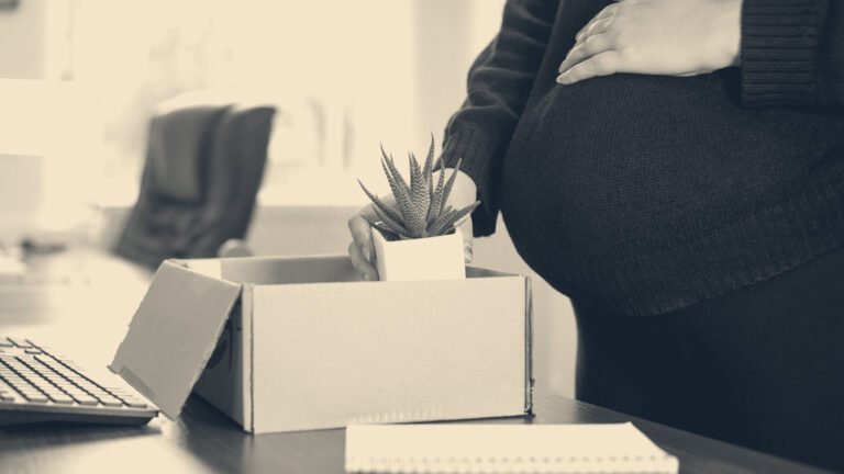 Pregnant woman cradling her bump packs a desk plant into a cardboard box in a bright office