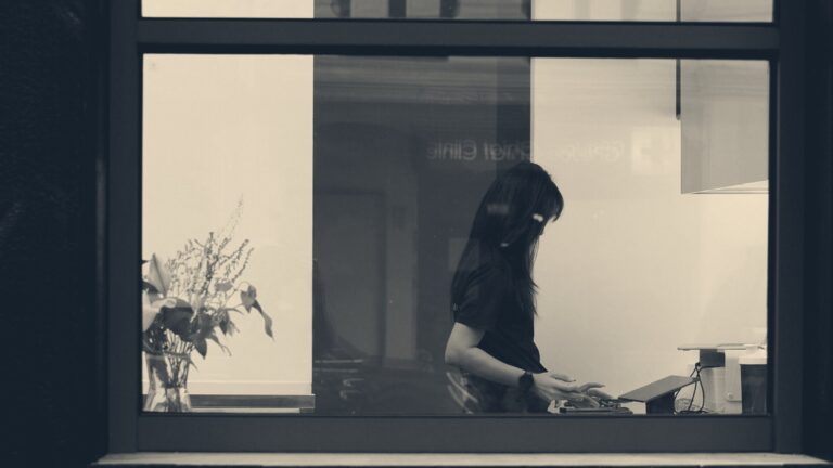 A woman working at a table inside a warm-lit room, seen alone through dark-framed office windows.