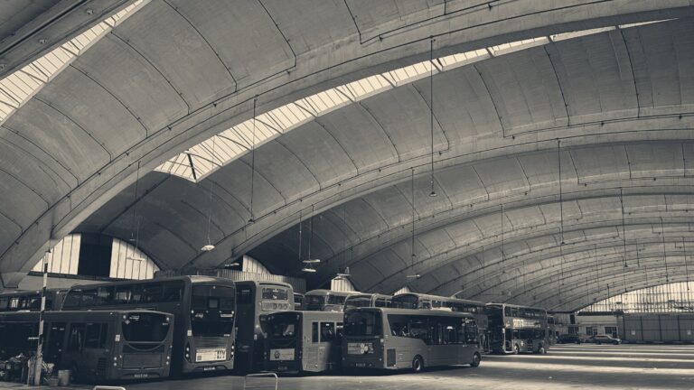 Red London buses parked beneath the soaring concrete cantilever arches of Stockwell Bus Garage.