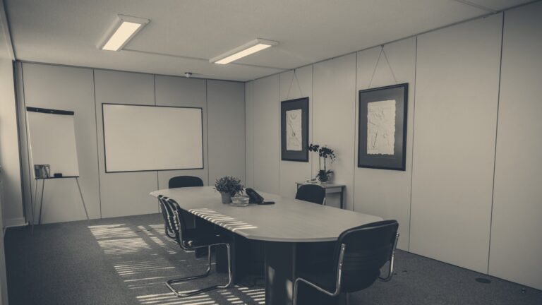 Empty UK office boardroom with conference table, chairs, whiteboard, and sunlight slanting through window blinds.