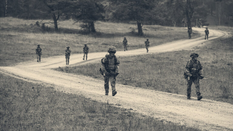 British officer cadets in camouflage march in file along a winding gravel track through an open heathland training area