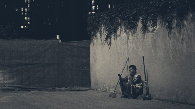 A night-shift cleaner in a hi-vis vest rests against a concrete wall, broom and dustpan beside him, lit tower windows behind