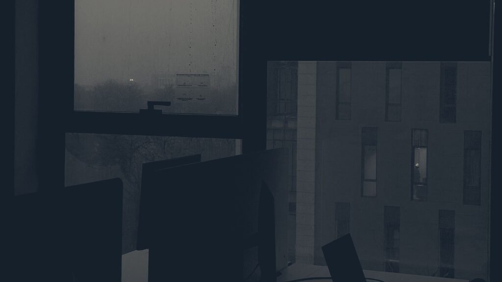 Dark office after hours: silhouetted monitors and laptop beside rain-streaked windows overlooking buildings at dusk