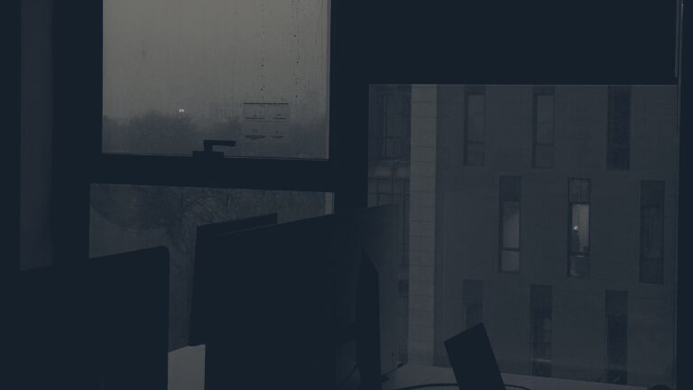 Dark office after hours: silhouetted monitors and laptop beside rain-streaked windows overlooking buildings at dusk