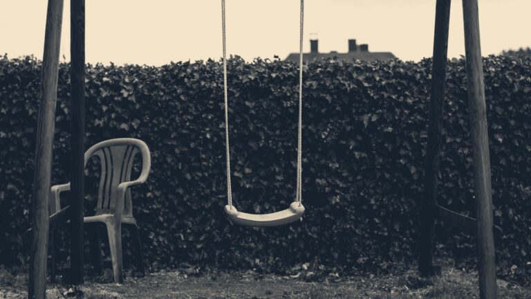 An empty pink swing seat hangs motionless in a garden before an ivy hedge and grey sky.