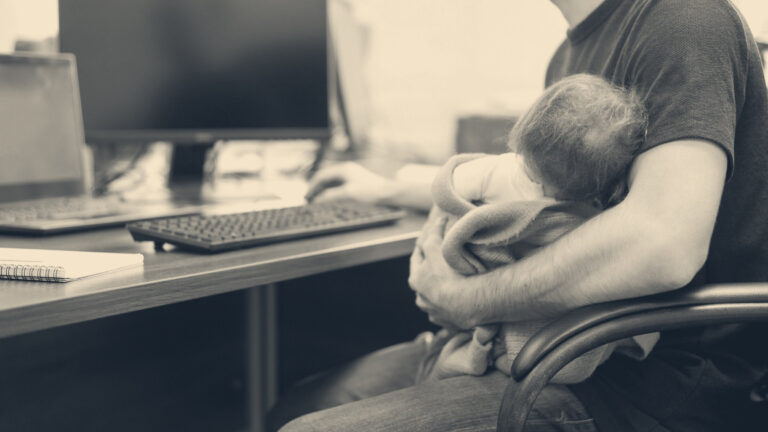 A father sitting at an office desk cradles a sleeping newborn baby wrapped in a blanket while working at a computer