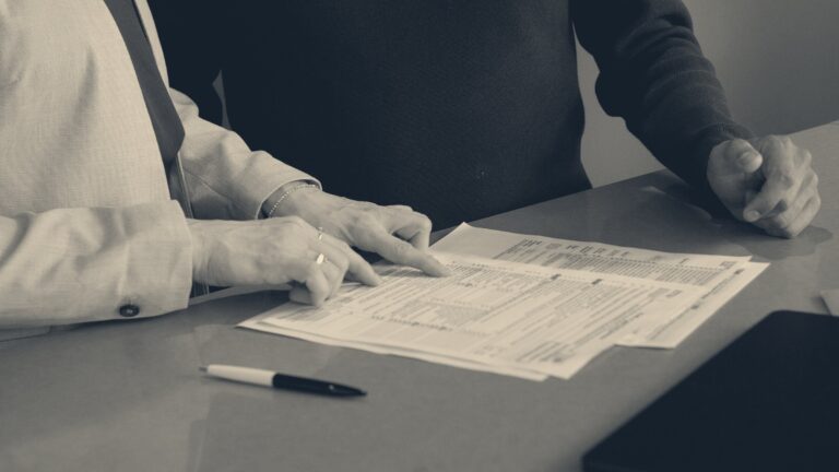Two colleagues reviewing performance documents across a meeting desk, one pointing at printed figures beside a pen.
