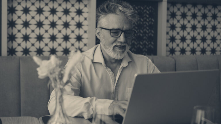 Grey-haired man with glasses typing on a laptop at a cafe table, a glass vase in the foreground.