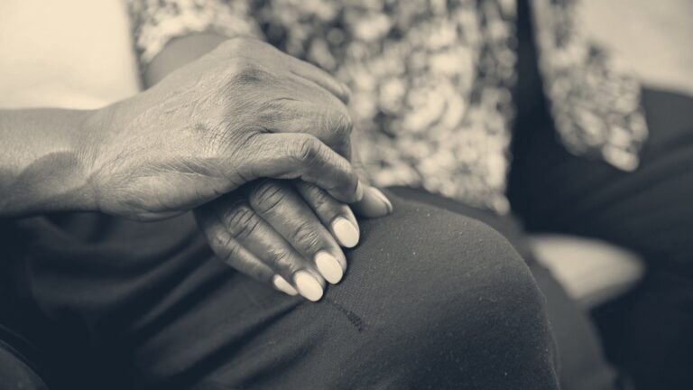 A caregiver gently holds an older person's weathered hand resting together on dark fabric on their lap.