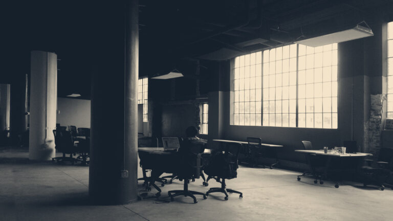 A lone worker at a laptop in a dim loft office, desks and chairs receding toward tall grid windows.