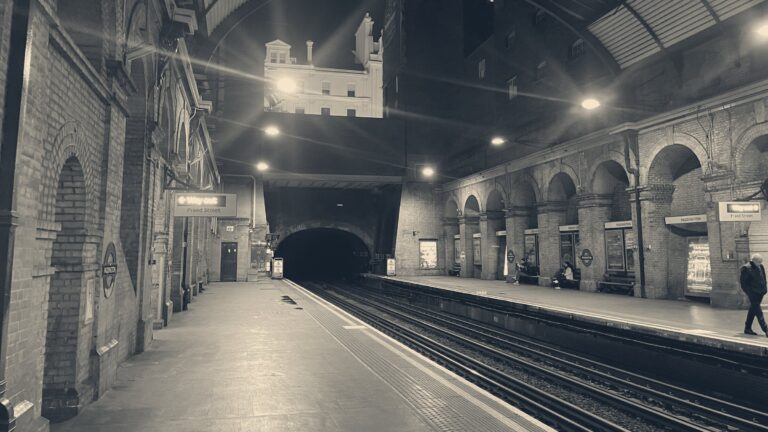 Near-empty London Underground platform at night under Victorian brick arches, facing a dark tunnel mouth.